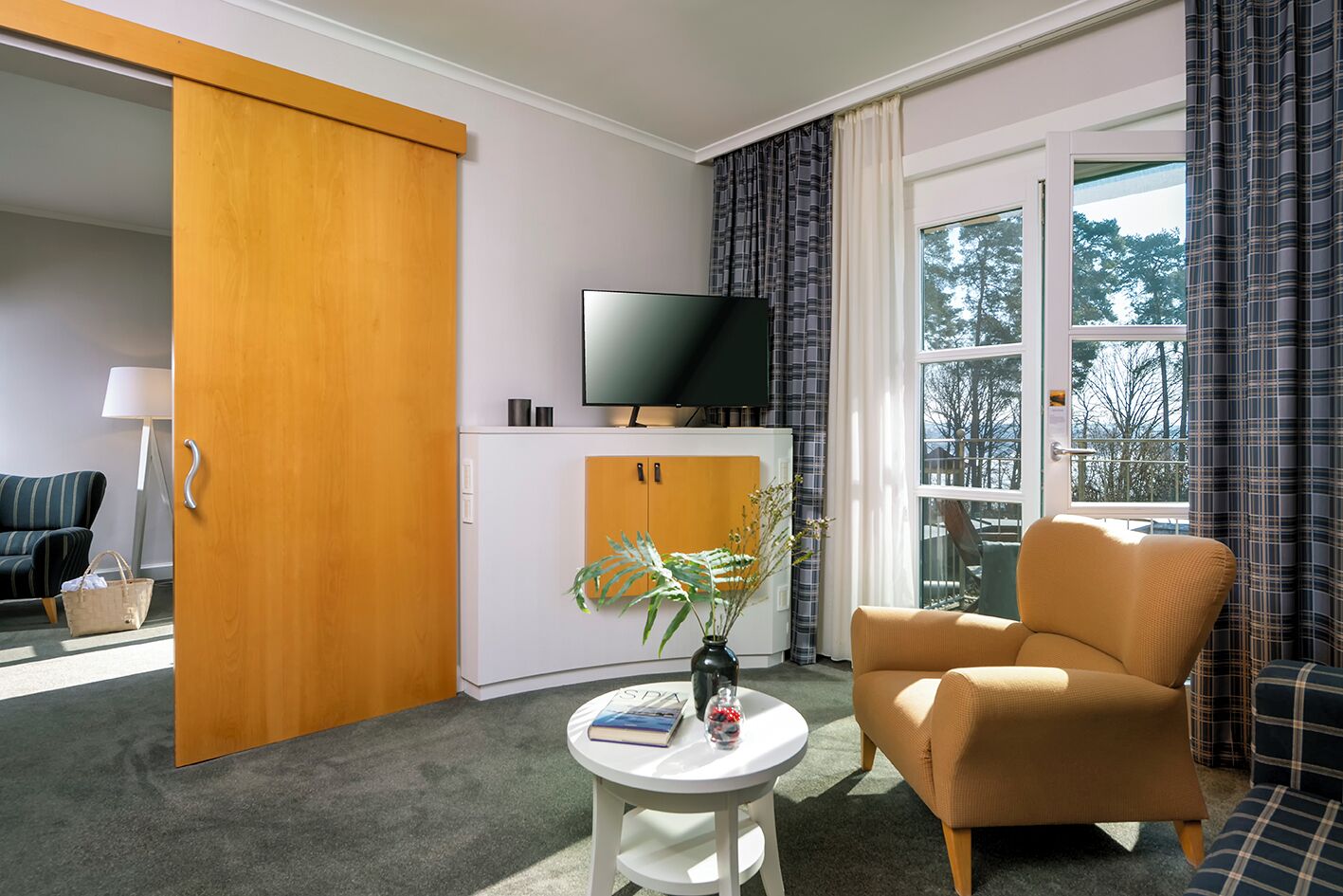 Ein Hotelzimmer mit Schiebetür, einem Fernseher auf einem Schrank, einem Sofa, einem kleinen Tisch mit Vase und Buch sowie Blick durch ein Fenster.