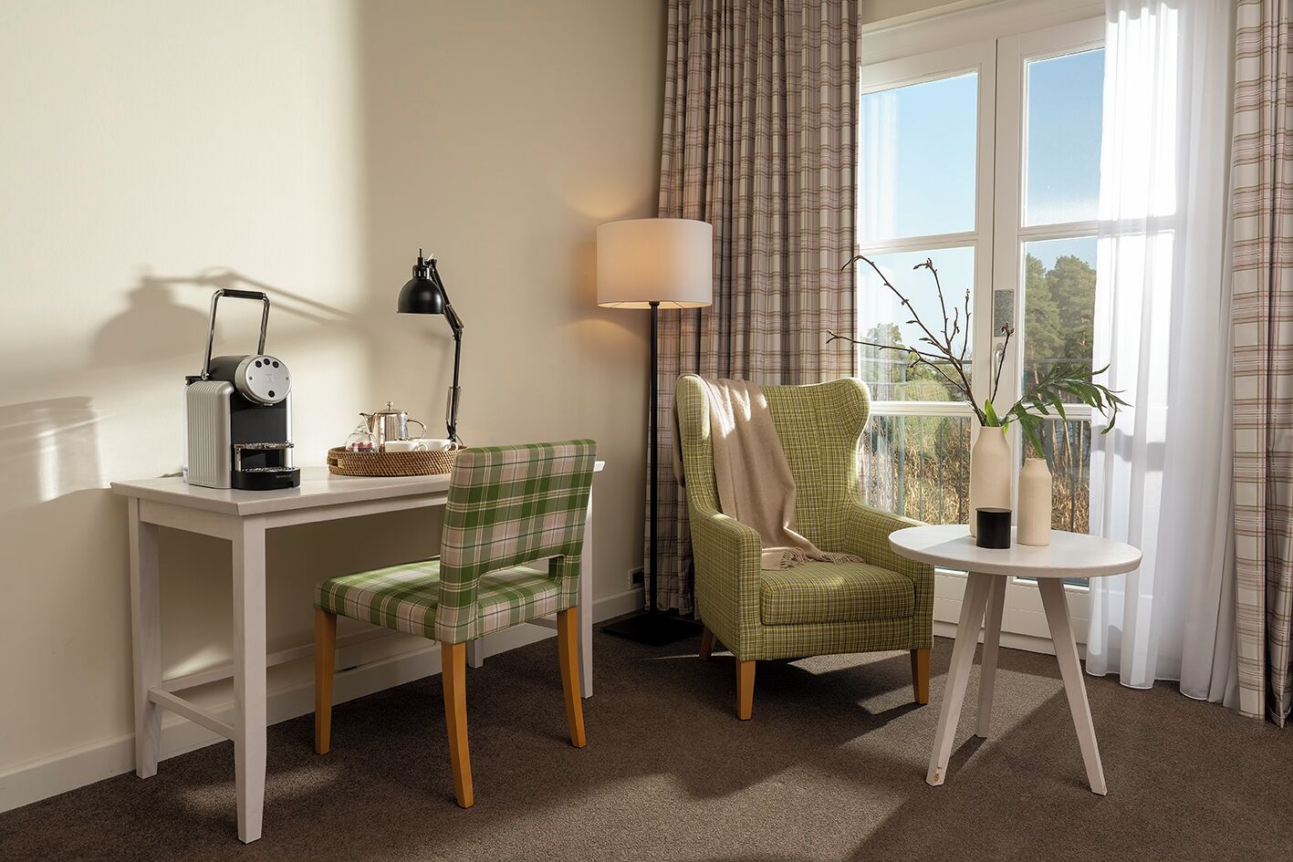 Une pièce avec un bureau blanc, une chaise à carreaux verts, une lampe et une vue sur l’extérieur par une fenêtre ornée de rideaux.