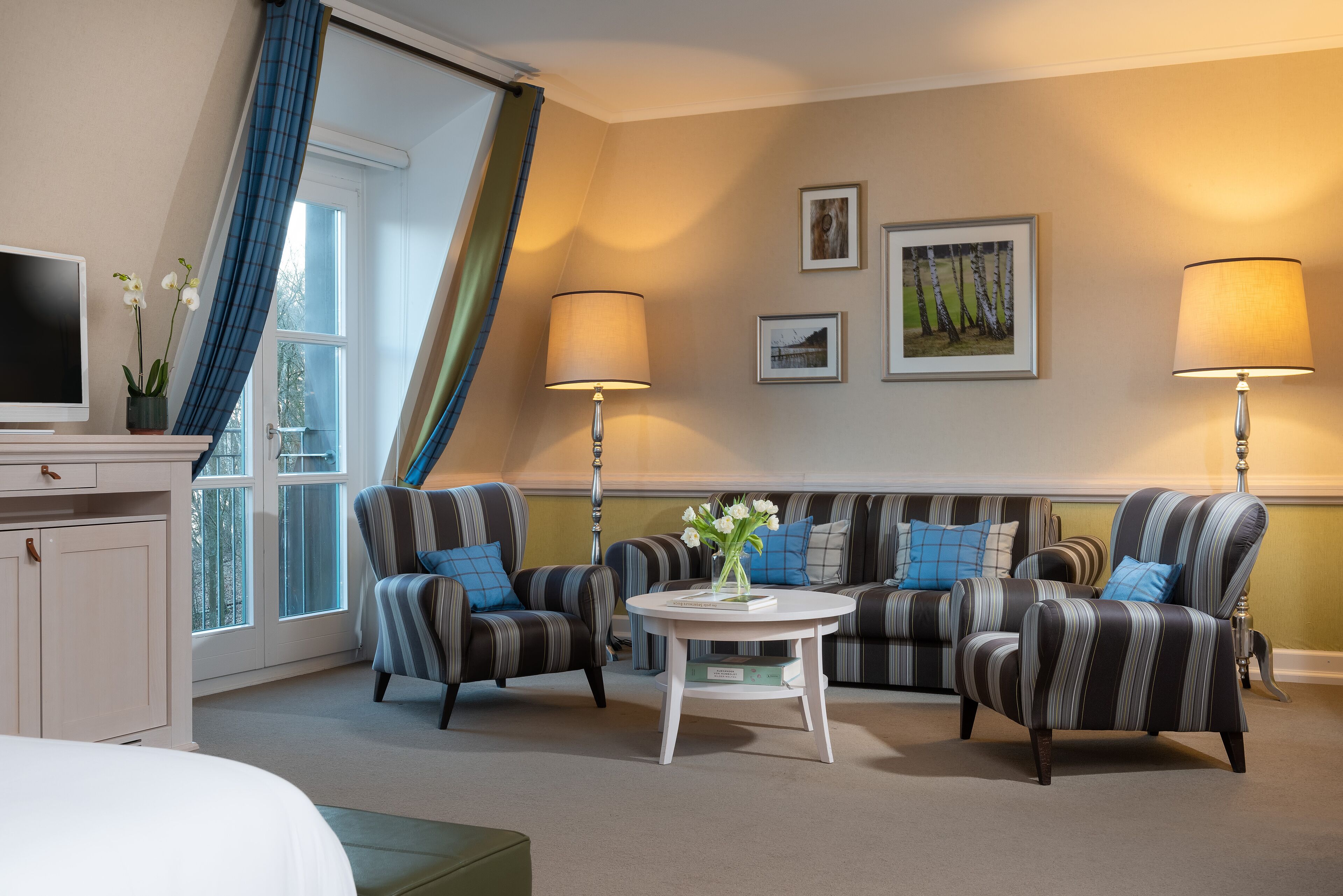 A hotel room with a sofa set, two armchairs, a small table with a vase of flowers, a TV, and a window with curtains.