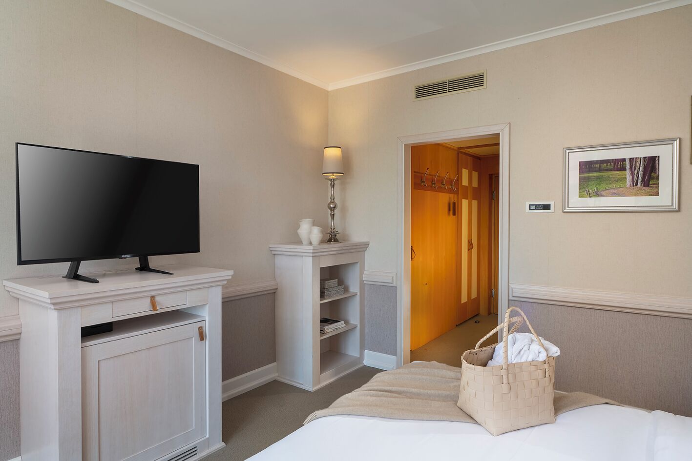 Habitación de hotel con televisión de pantalla plana sobre un mueble blanco, una lámpara de noche, una cama con manta color beige, una cesta de mimbre y un cuadro en la pared.