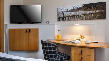 Et hotelværelse med en seng, et fjernsyn ophængt på væggen, et træskrivebord med stol, et billede og et sengebord med lampe og kop.