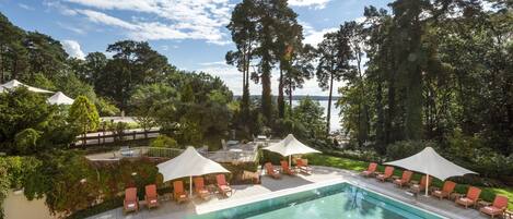 A swimming pool with lounge chairs and umbrellas, surrounded by tall trees and a clear sky.