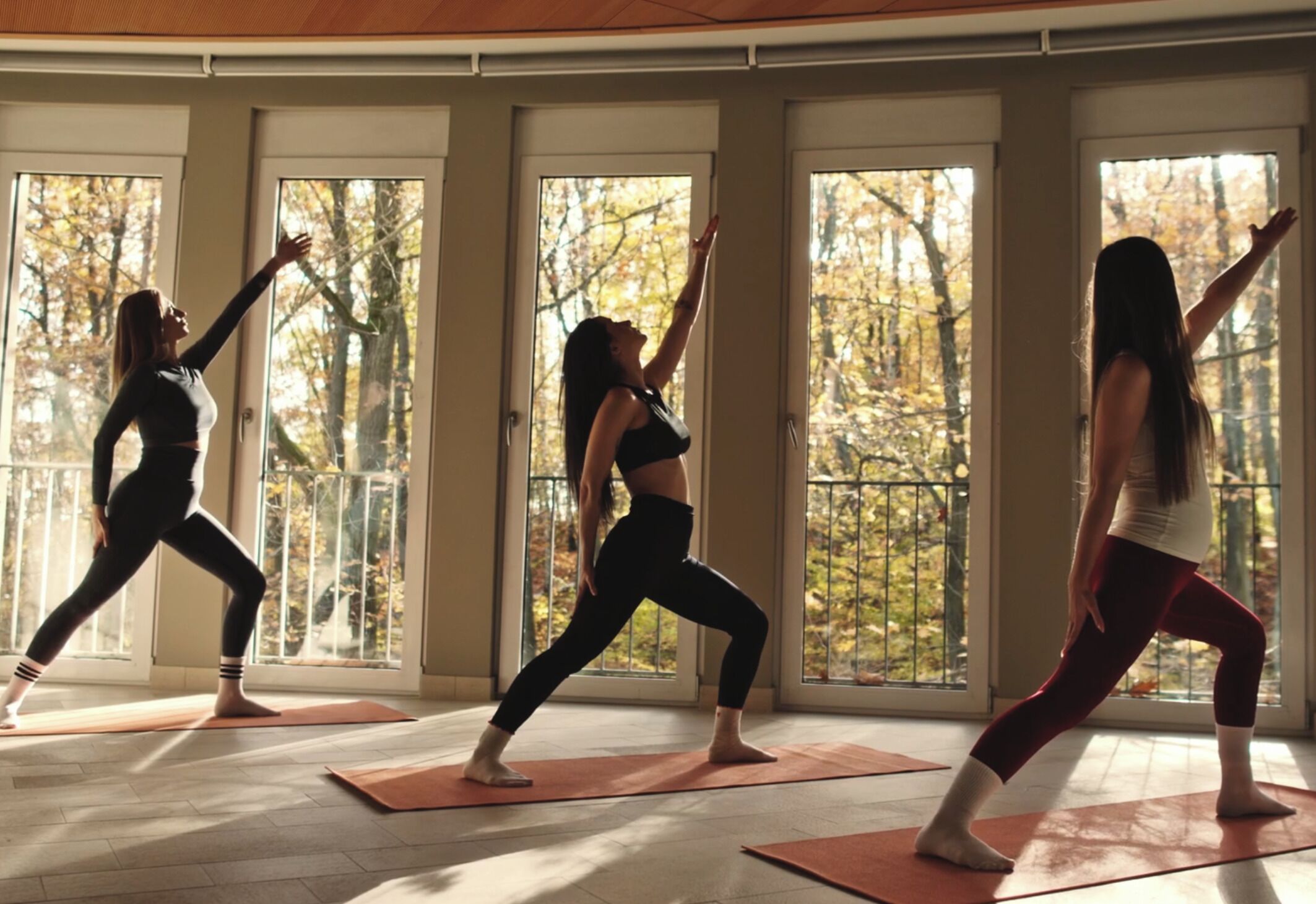 Tres personas practicando yoga en una habitación con ventanas amplias y vistas a árboles.