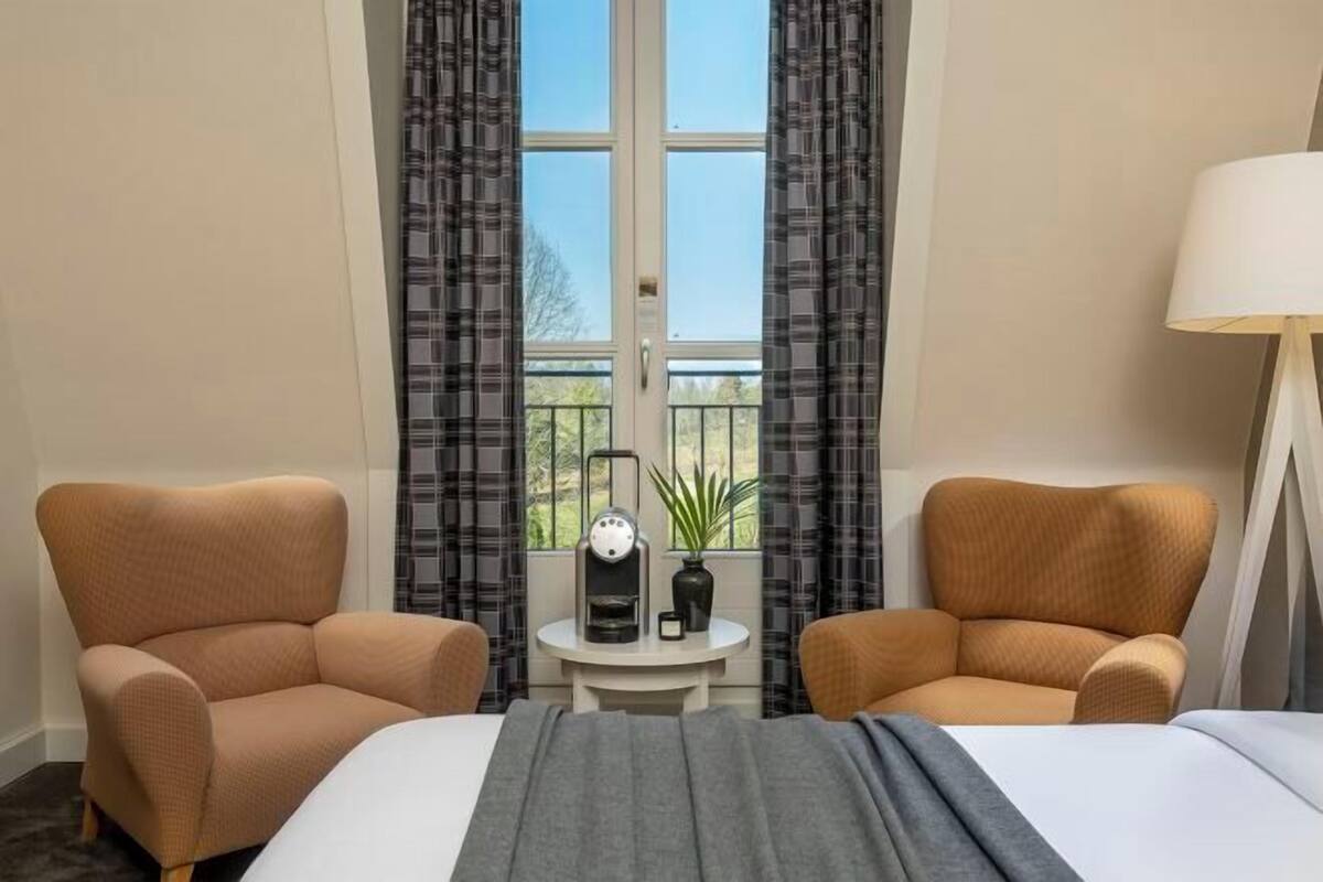 Una habitación de hotel con una cama, dos sillones, una mesita con cafetera y vistas al exterior a través de una ventana.