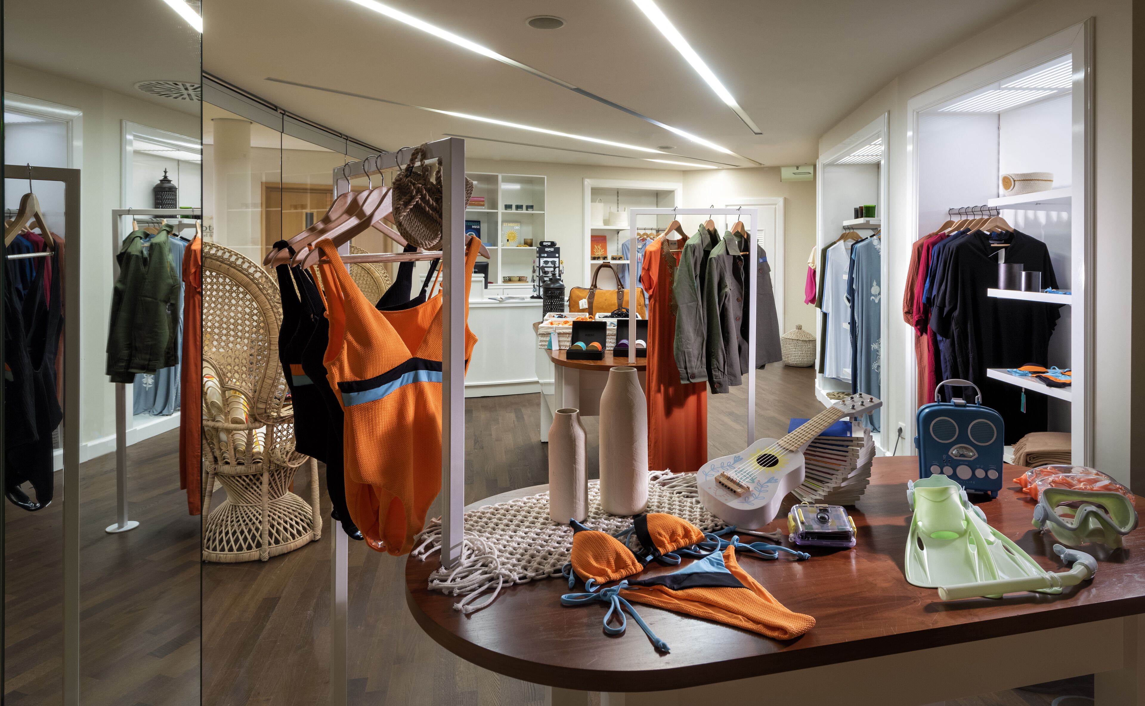 A well-lit retail space with clothing racks, a display table with various items, and a wicker chair.
