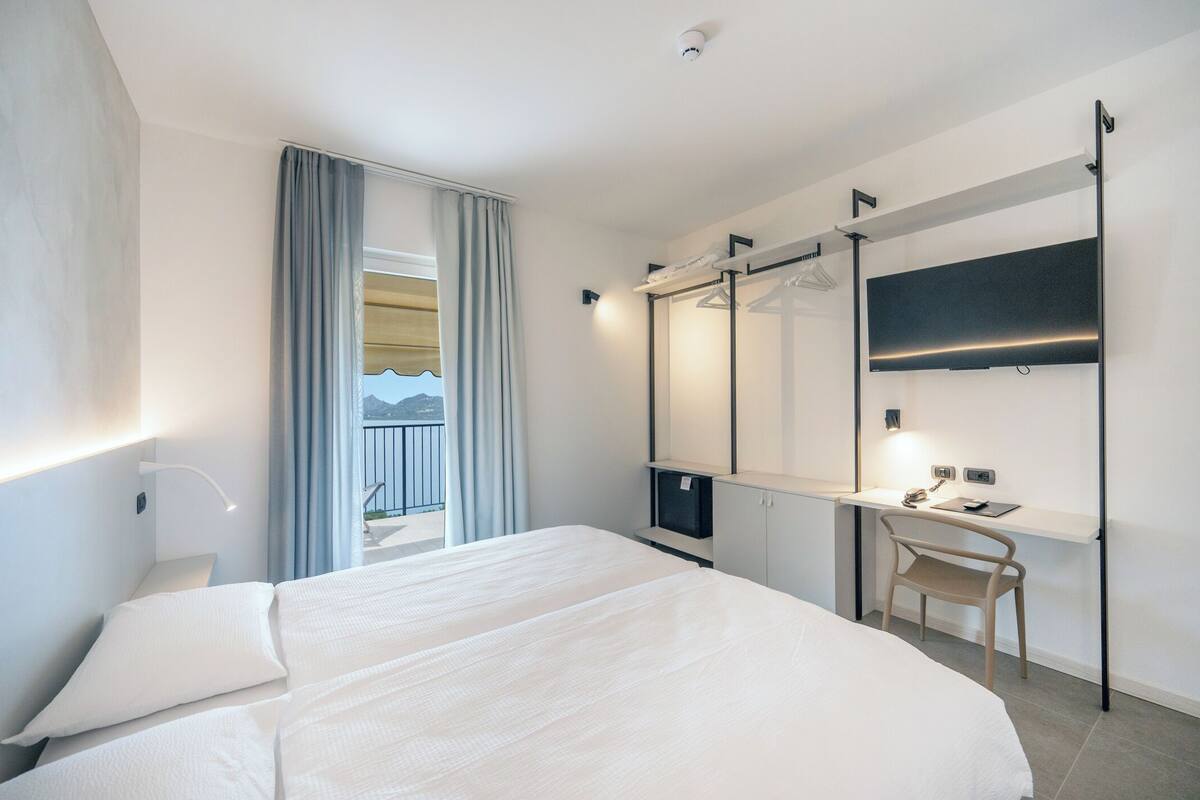 A modern hotel room with a large bed, a desk with a chair, a flat-screen TV, and a balcony with curtains.