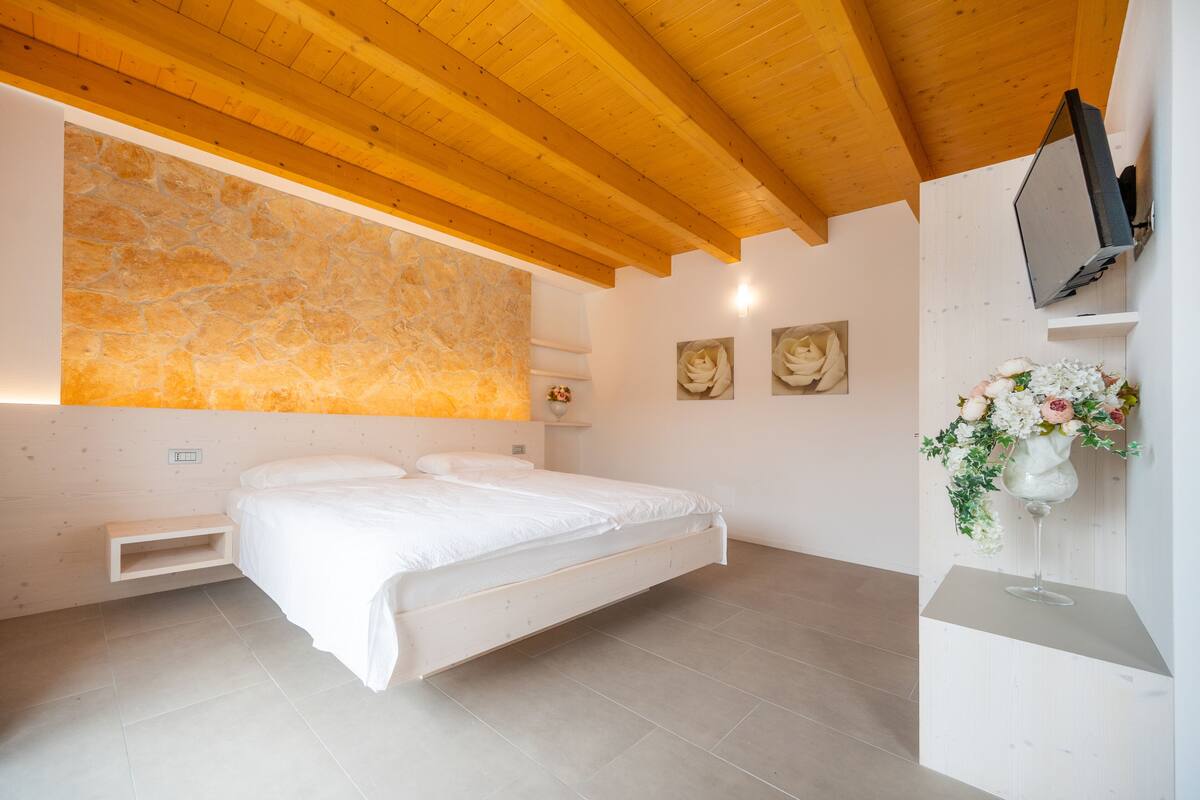 A bedroom with a bed, a stone wall, wooden ceiling beams, a TV, and a flower arrangement.