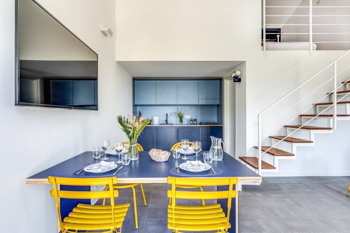 Ein moderner Essbereich mit einem dunkelblauen Tisch und gelben Stühlen, eine Küche mit blauen Schränken und eine Treppe mit Holzboden.
