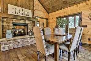 A wooden cabin interior with a stone fireplace, four upholstered chairs with metal studs, a wooden dining table, and a large window with a view of greenery.