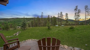 Een terras met twee Adirondack-stoelen en een tafel, met uitzicht op een gazon met bomen en een bergketen in de verte.