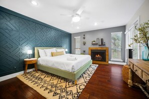 Une chambre moderne avec un lit vert, une table de chevet en bois, un tapis à motifs, une chemin ée et une vue sur l’extérieur grâce à de grandes fenêtres.