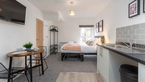 Habitación de hotel compacta con cama, kitchenette pequeña, comedor con mesa y sillas, y televisor montado en la pared.