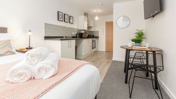 Una habitación de hotel moderna con una cama bien hecha, una pequeña kitchenette, una mesa redonda con sillas y un televisor montado en la pared.