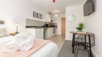 Una habitación de hotel moderna con una cama bien hecha, una pequeña kitchenette, una mesa redonda con sillas y un televisor montado en la pared.