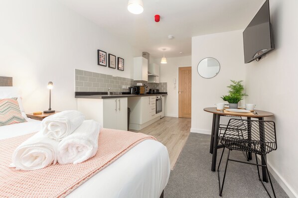 A modern hotel room with a neatly made bed, a small kitchenette, a round dining table with chairs, and a TV mounted on the wall.