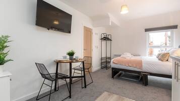 Una habitación de hotel moderna con una cama, un televisor montado en la pared, una pequeña mesa de comedor con sillas, una planta en maceta y una ventana con vistas.