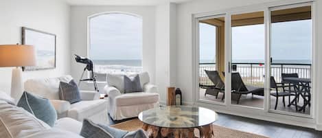 A spacious living room with a large window overlooking the beach, a ceiling fan, and a round glass coffee table.