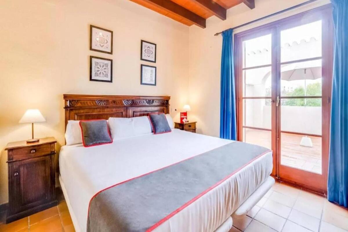 Une chambre d’hôtel avec un grand lit, une tête de lit en bois, deux tables de chevet avec des lampes et un balcon agrémenté de rideaux bleus.