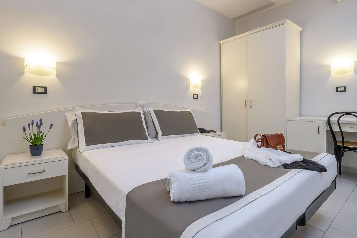 A neatly made bed with a rolled towel, a small bedside table with a plant, and a wardrobe in a hotel room.