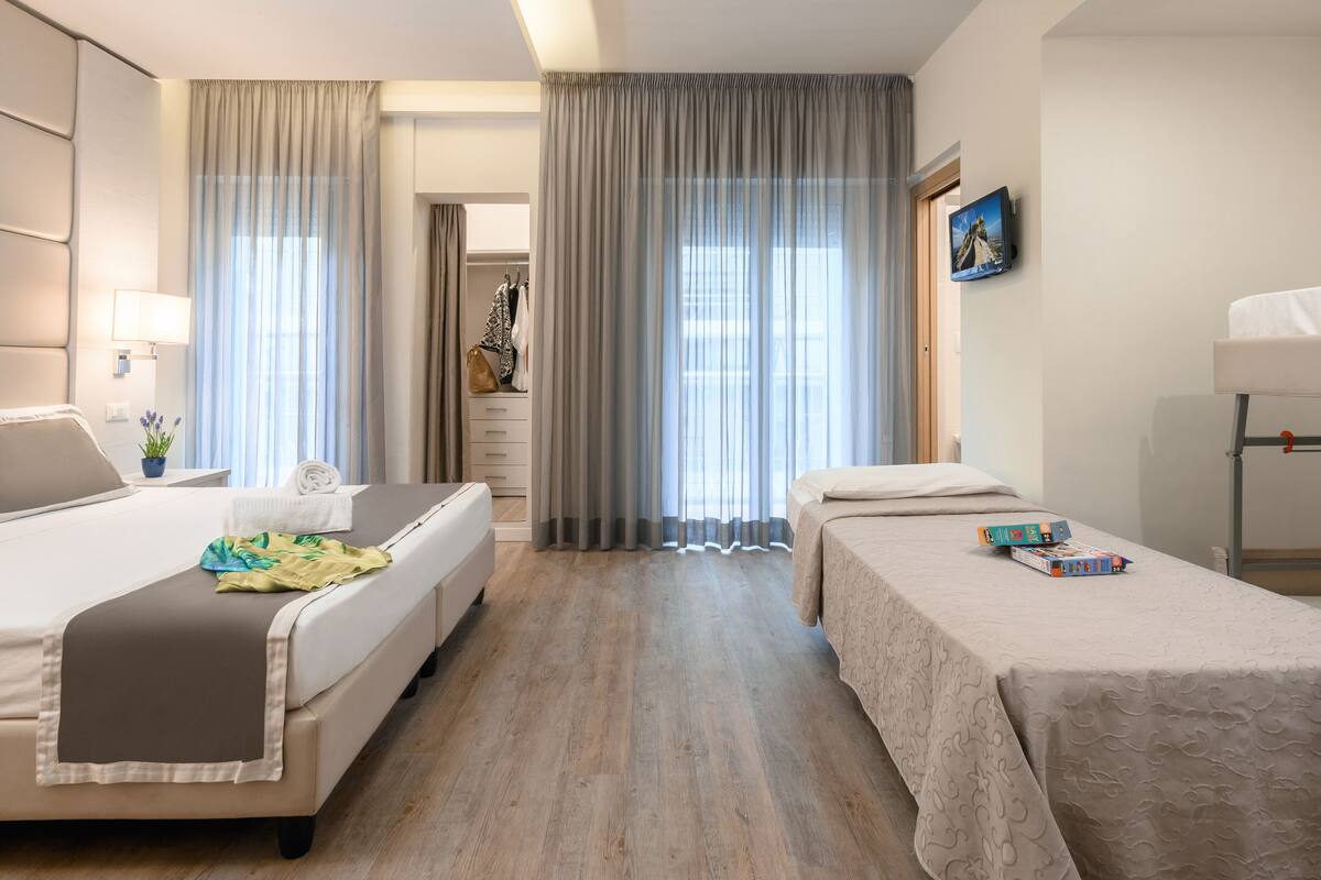 A hotel room with two beds, a bedside table with a lamp, a dresser, and a large window with curtains.