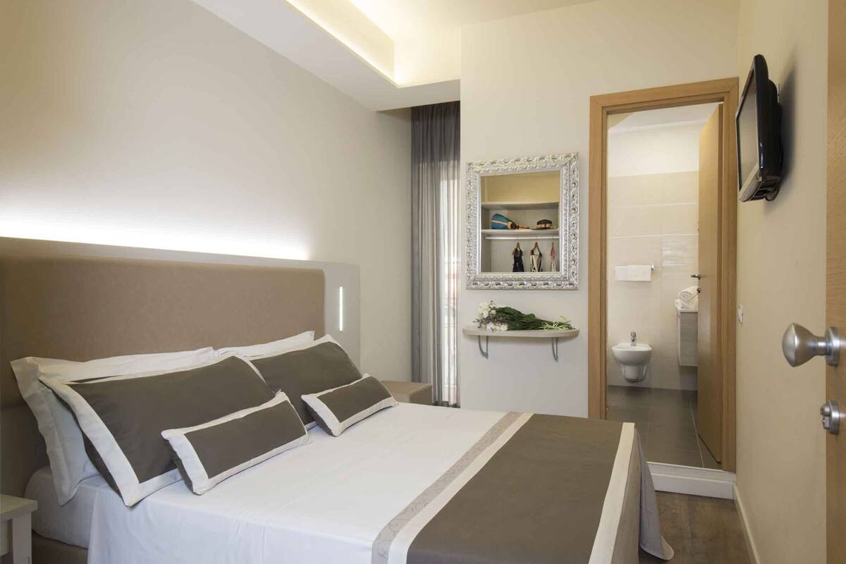 A modern hotel room with a large bed, a bedside table, a mirror, and a bathroom visible through an open door.