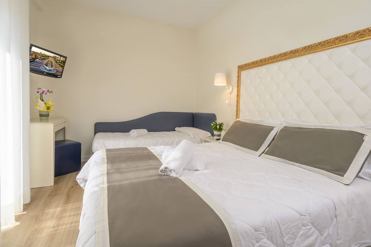 A hotel room with two beds, a TV mounted on the wall, a bedside table with a vase of flowers, and a wall-mounted lamp.