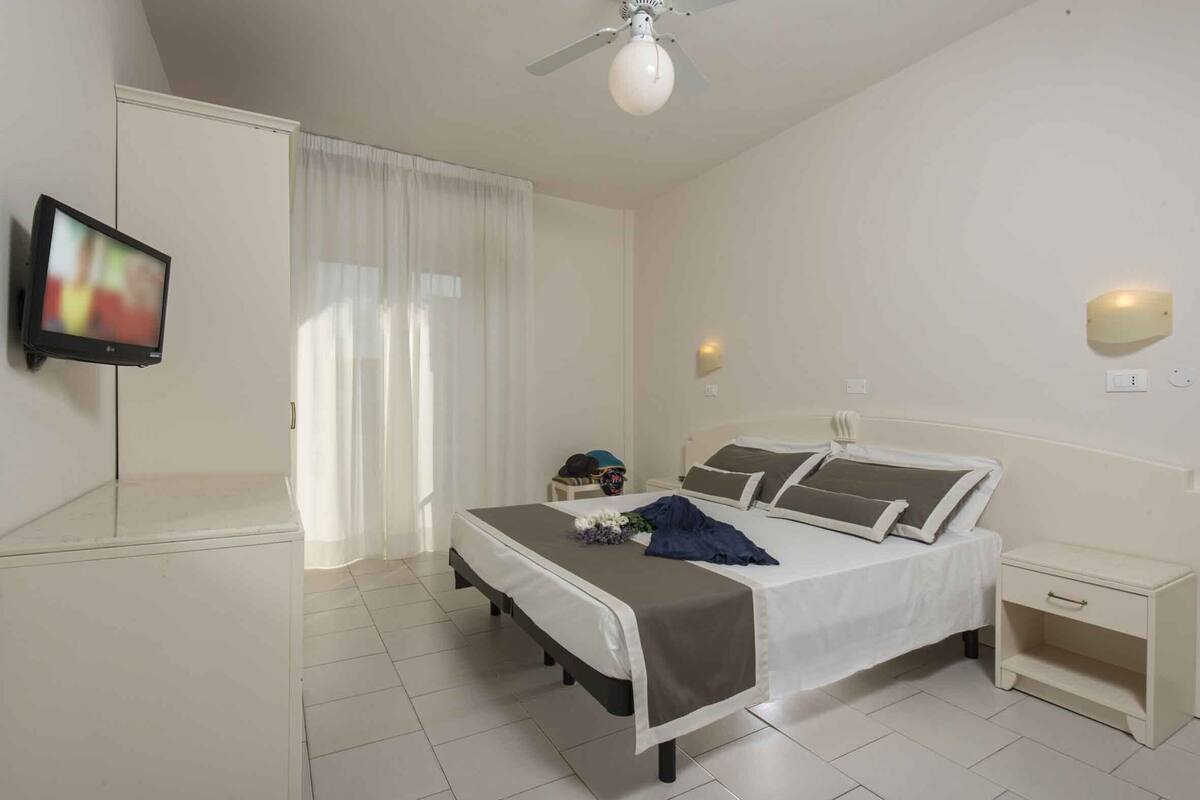 A bedroom with a bed, bedside table, nightstand, ceiling fan, and a television mounted on the wall.