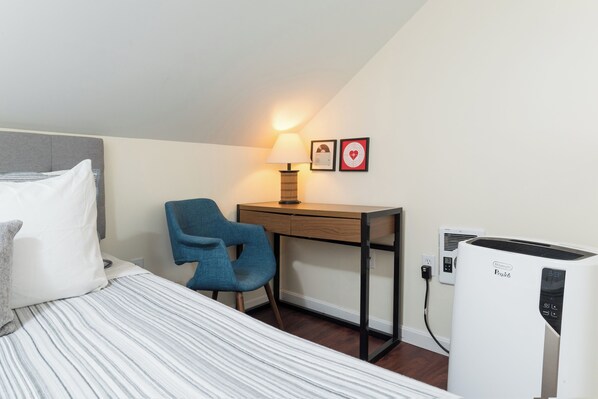 A hotel room with a bed, a desk, a chair, a lamp, two framed pictures, and an air conditioner.