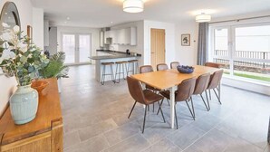 A modern dining area with a wooden table and chairs, a kitchen in the background, and a balcony with a railing.