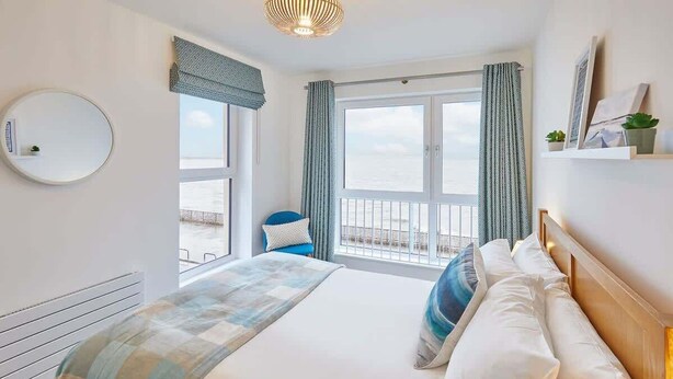 A bedroom with a large bed, a round mirror, a view of the sea, and a balcony with a chair.
