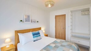 A neatly arranged bedroom with a wooden bed, a bedside table with a lamp, a wardrobe, and a shelf with a framed quote.