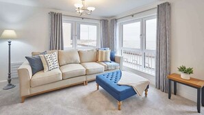 A living room with a beige sofa, a blue ottoman, a floor lamp, and a small table with a plant.