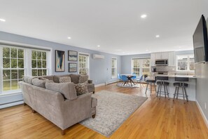 Un salon spacieux avec un grand canapé beige, un coin repas avec des chaises bleues et une cuisine dotée de meubles blancs et d’une grande fenêtre.