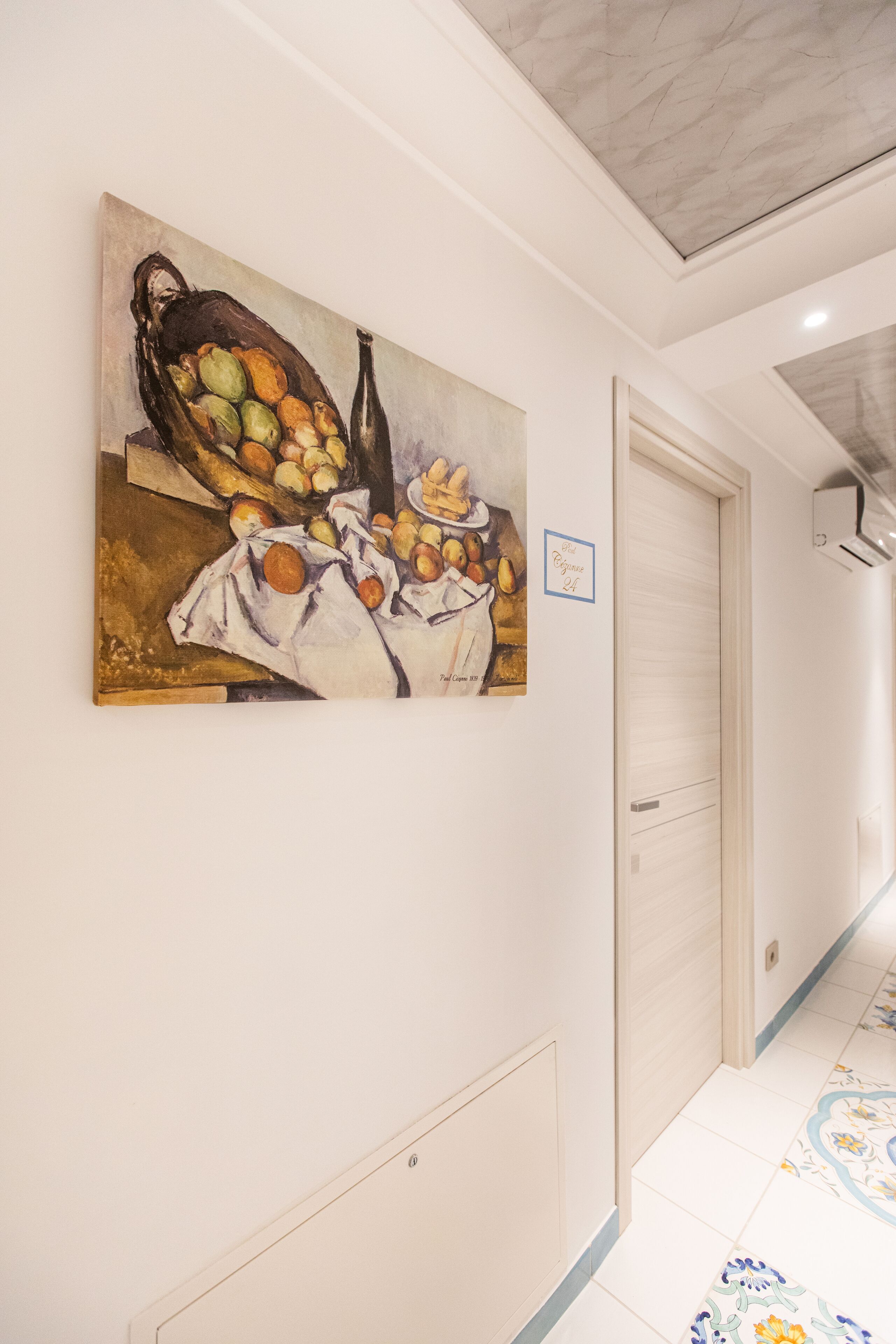 Un pasillo con un cuadro de frutas y verduras en la pared, una puerta cerrada y baldosas de suelo con diseños.