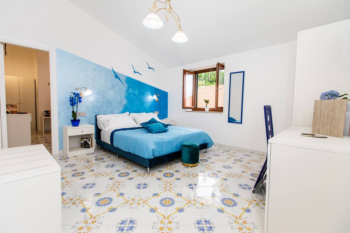 A modern bedroom with a blue and white color scheme, a bed with a blue headboard, a small bedside table with a potted plant, and a window with a view of greenery outside.