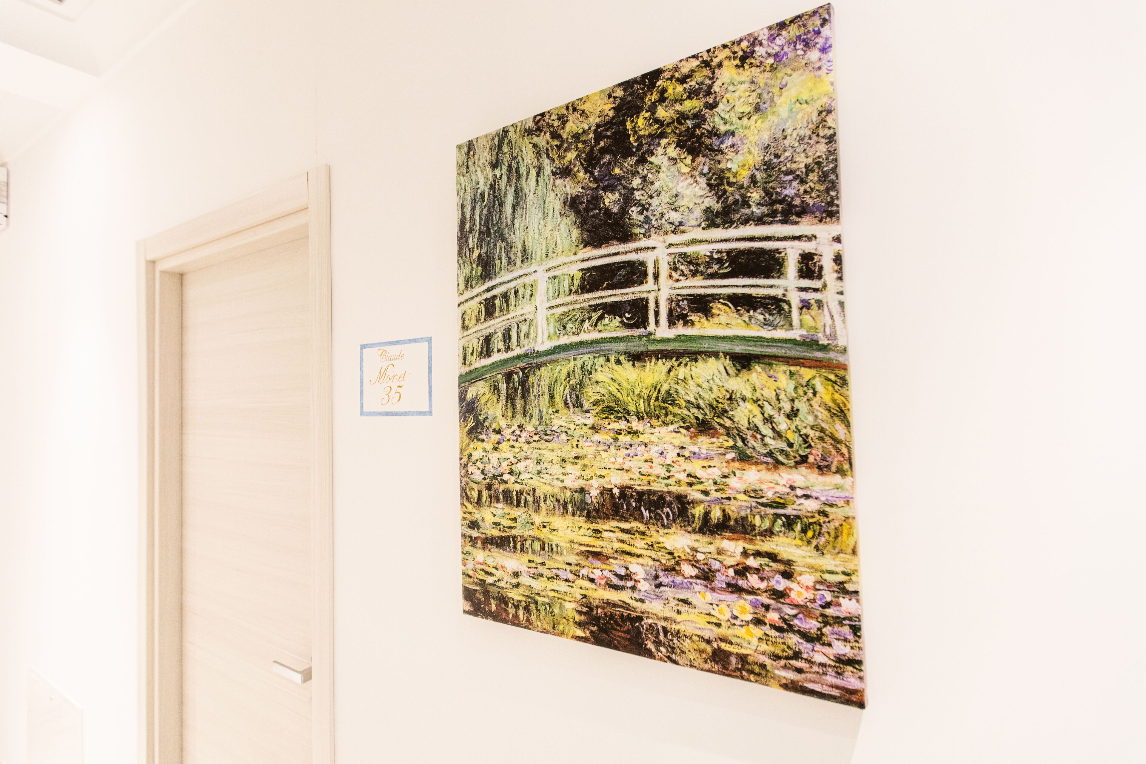 A room with a white door, a framed painting of a bridge over water, and a small sign on the wall.