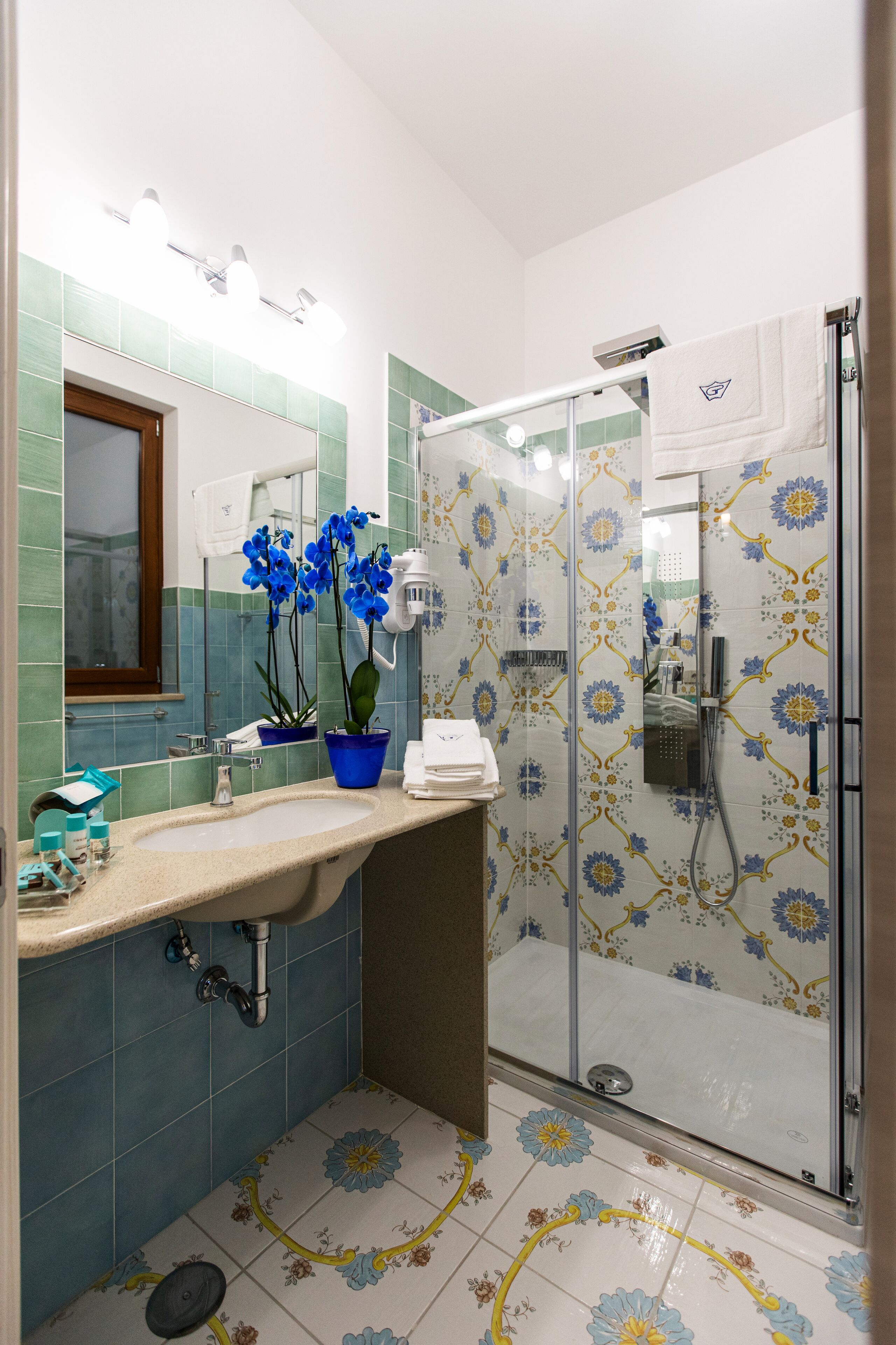A bathroom with a sink, mirror, blue orchid plant, shower, and patterned tiles.
