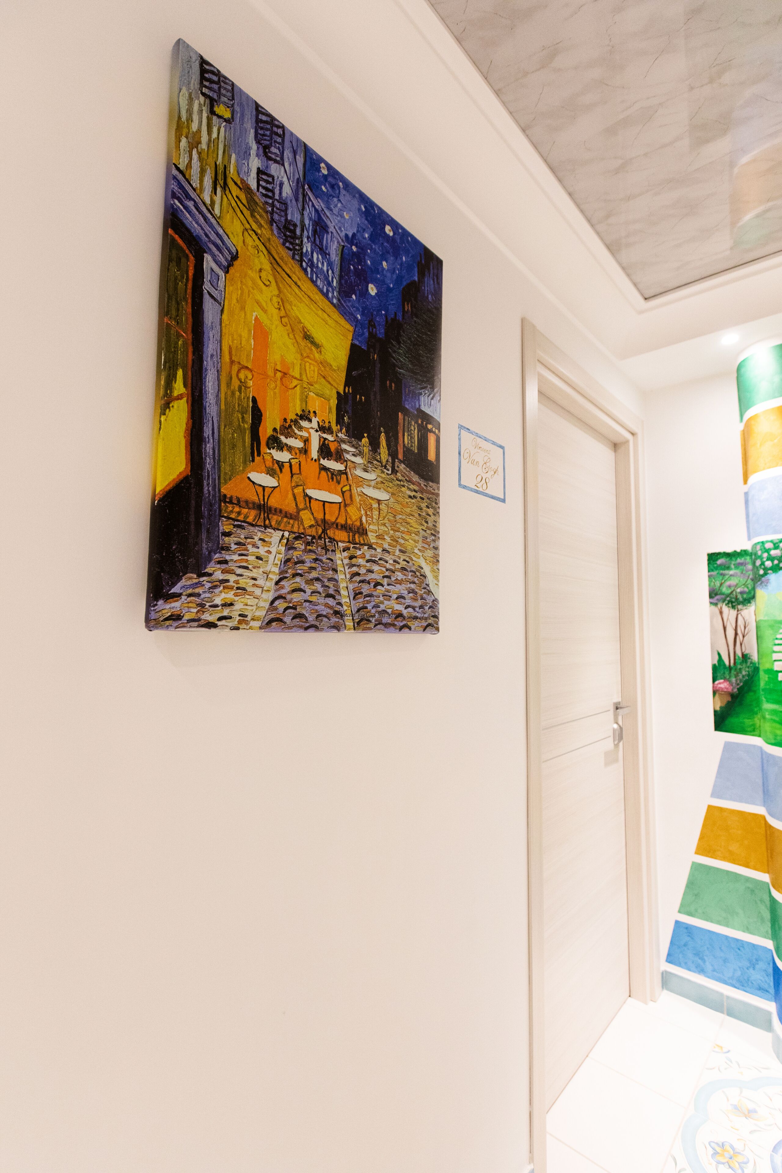 Un pasillo con un cuadro que muestra una escena de café, una puerta y una pared con rayas de colores.