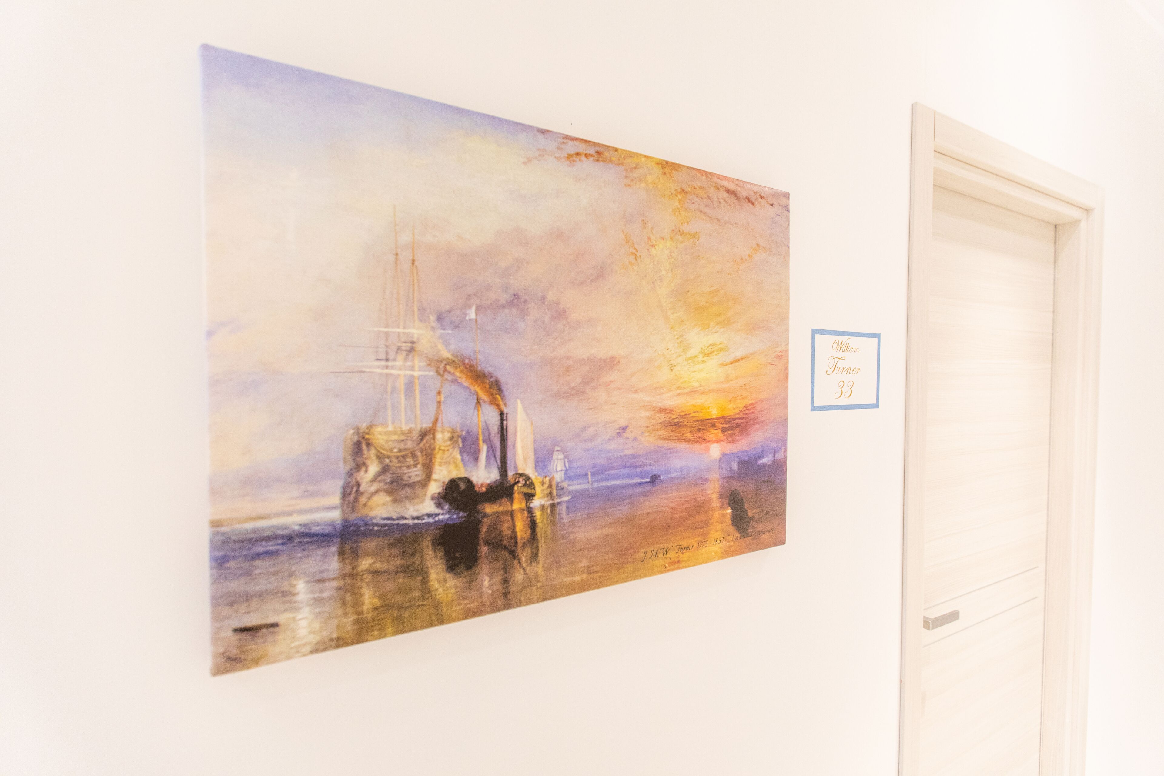 En målning som föreställer en solnedgång över ett lugnt hav med fartyg, upphängd på en vit vägg.