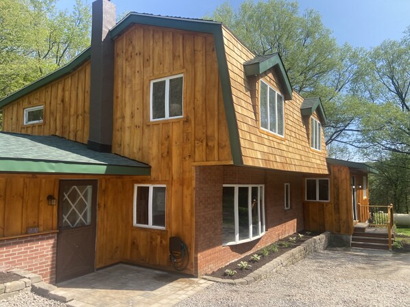 A two-story wooden house with a green roof, brick foundation, and a gravel driveway.