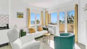 Una stanza luminosa e soleggiata con un divano bianco, una poltrona verde acqua e un piccolo tavolo rotondo. Ampie finestre offrono vista sul mare e un balcone con mobili da esterno.