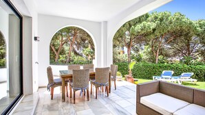 A covered patio with wicker furniture, a glass table, and a view of a garden with trees and a hedge. - N07 Cabopino (Marbella)
