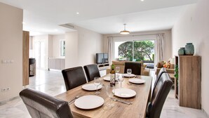 A dining area with a wooden table set for four, surrounded by leather chairs. A television is visible in the background, and a window offers a view of greenery outside. - N07 Cabopino (Marbella)