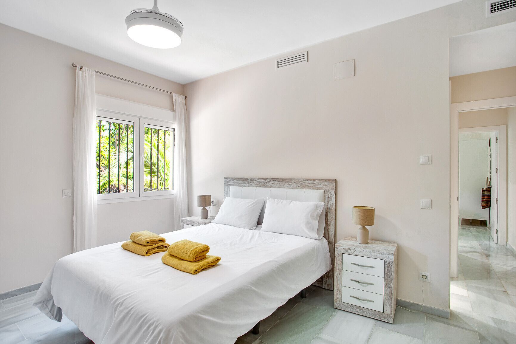 A bedroom with a bed, two bedside tables, a window with curtains, and a view of greenery outside.