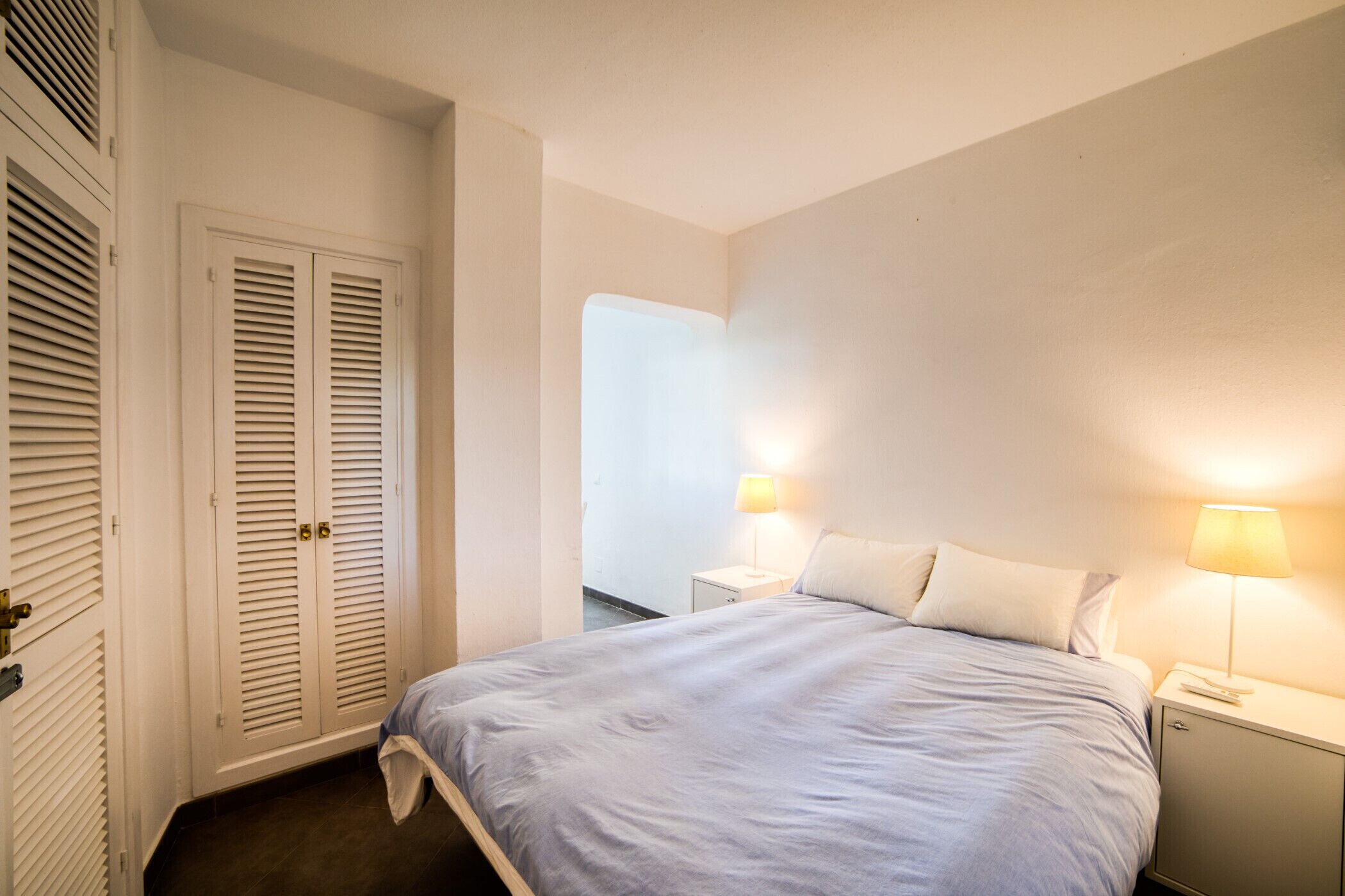 A bedroom with a bed, two bedside lamps, a nightstand, and a wardrobe with louvered doors.