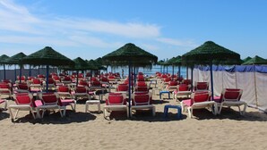 Una spiaggia con file di sdraio rosse e bianche sotto ombrelloni di paglia, sabbia dorata e cielo azzurro limpido.