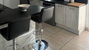 Una cocina moderna con una mesa negra para comer, dos taburetes de bar, un refrigerador, un fregadero y una ventana con persianas.