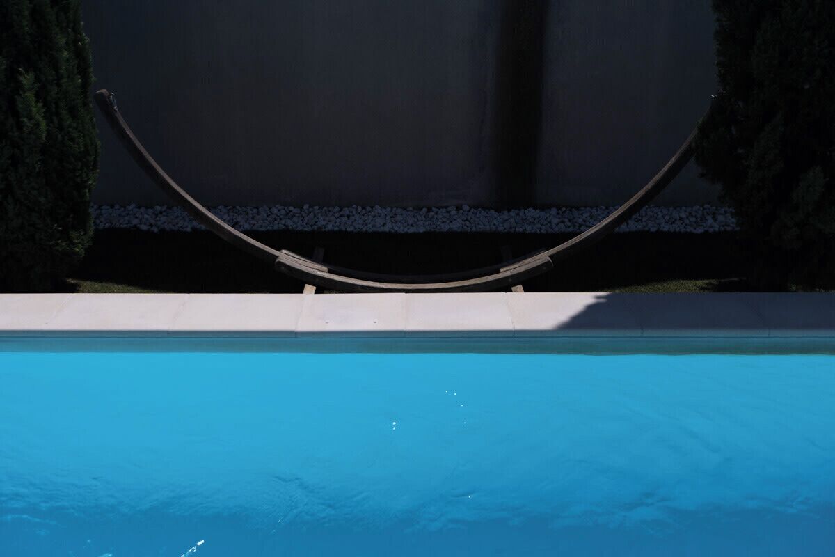 A swimming pool with a curved structure above it, surrounded by a low wall and greenery.