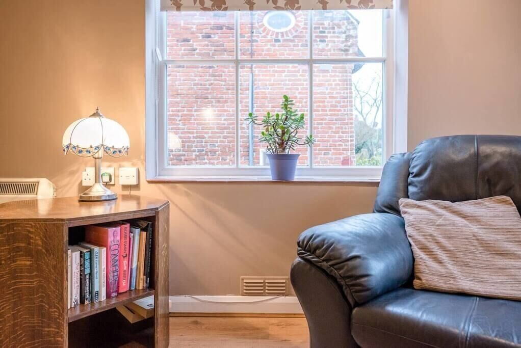 A room with a leather sofa, a bookshelf with books, a lamp, and a window with a brick wall view.