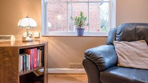 A room with a leather sofa, a bookshelf with books, a lamp, and a window with a brick wall view.